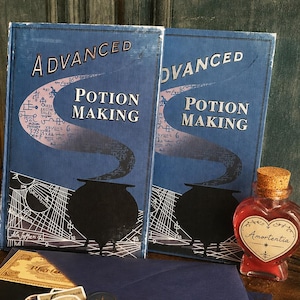 May include: Two blue "Advanced Potion Making" books with a cauldron illustration. A heart-shaped bottle of red liquid labeled "Amortentia" sits next to the books. A dark blue envelope with a wax seal and stickers is also present.