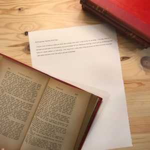 May include: A close-up of an open book with a white page showing a typed letter. The letter is about a woman who is staying with a friend while her husband is away. The book is on a wooden table with a stack of books in the background.