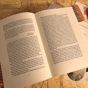 K&ouml;nnte beinhalten: Ein aufgeschlagenes Buch mit wei&szlig;en Seiten und schwarzem Drucktext, das die Seiten 14, 15 und 16 des Romans Dracula zeigt. Das Buch liegt auf einer Oberfl&auml;che mit einer alten Karte von London und weiteren Papierbeilagen, darunter ein Ticket.