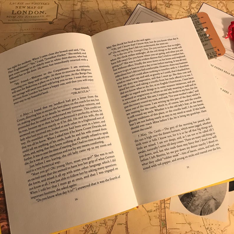 K&ouml;nnte beinhalten: Ein aufgeschlagenes Buch mit wei&szlig;en Seiten und schwarzem Drucktext, das die Seiten 14, 15 und 16 des Romans Dracula zeigt. Das Buch liegt auf einer Oberfl&auml;che mit einer alten Karte von London und weiteren Papierbeilagen, darunter ein Ticket.
