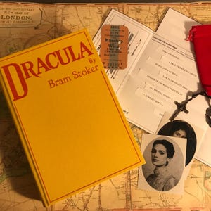 K&ouml;nnte beinhalten: Ein gelbes gebundenes Buch mit dem Titel Dracula von Bram Stoker, eine alte Fahrkarte von M&uuml;nchen nach Wien, ein bedruckter Brief mit Text, ein schwarzer Rosenkranz mit Kreuz, ein roter Stoffbeutel und ein Schwarz-Wei&szlig;-Portr&auml;tfoto einer Person.