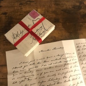 Puede incluir: Un montón de cartas antiguas atadas con una cinta roja. Las cartas tienen escritura manuscrita y un sello postal rosa. Una carta abierta revela más texto manuscrito. Las cartas están sobre una superficie de madera.