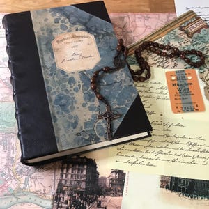 May include: A vintage journal with a blue marbled cover and black leather spine. A rosary with a crucifix lies across the journal. Other items include old maps, postcards, and a train ticket. The journal has the text "Smokins, Chompkins SOLICITORS".
