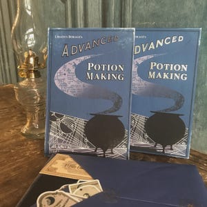 Puede incluir: Dos libros azules titulados Advanced Potion Making de Libatius Borage, con un gráfico de caldero negro y líneas geométricas blancas. Debajo de los libros hay un sobre azul oscuro que contiene varios recortes de papel pequeños y pegatinas.