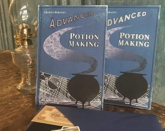 Puede incluir: Dos libros azules titulados Advanced Potion Making de Libatius Borage, con un gráfico de caldero negro y líneas geométricas blancas. Debajo de los libros hay un sobre azul oscuro que contiene varios recortes de papel pequeños y pegatinas.