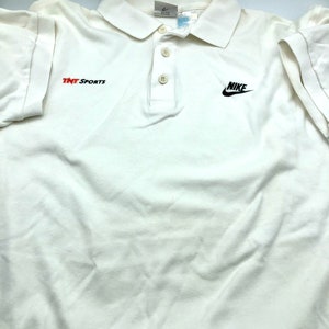 Pu&ograve; includere: Polo bianco con logo Nike nero e testo "TNT Sports" rosso sul davanti.