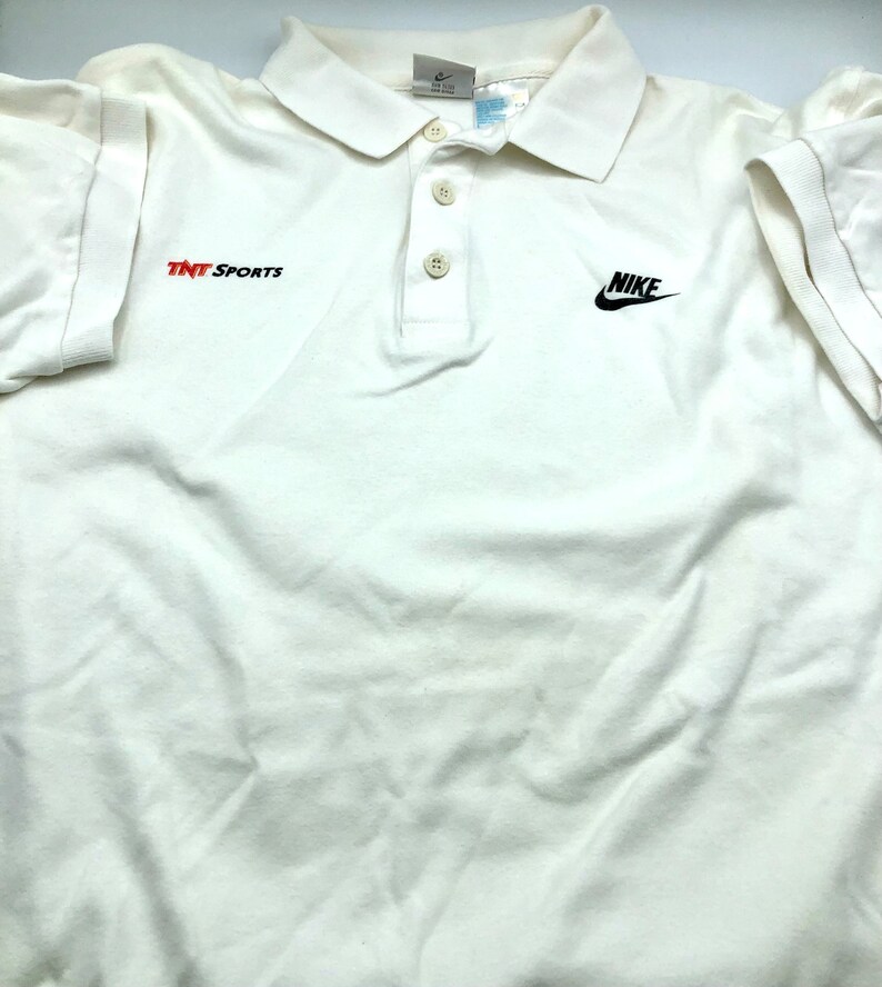 Pu&ograve; includere: Polo bianco con logo Nike nero e testo "TNT Sports" rosso sul davanti.