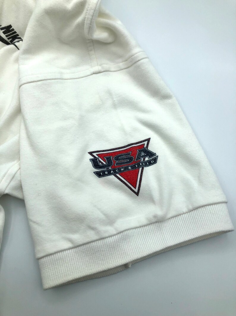 Pu&ograve; includere: Maglietta bianca a maniche corte con un logo USA Track & Field rosso, bianco e blu sulla manica.