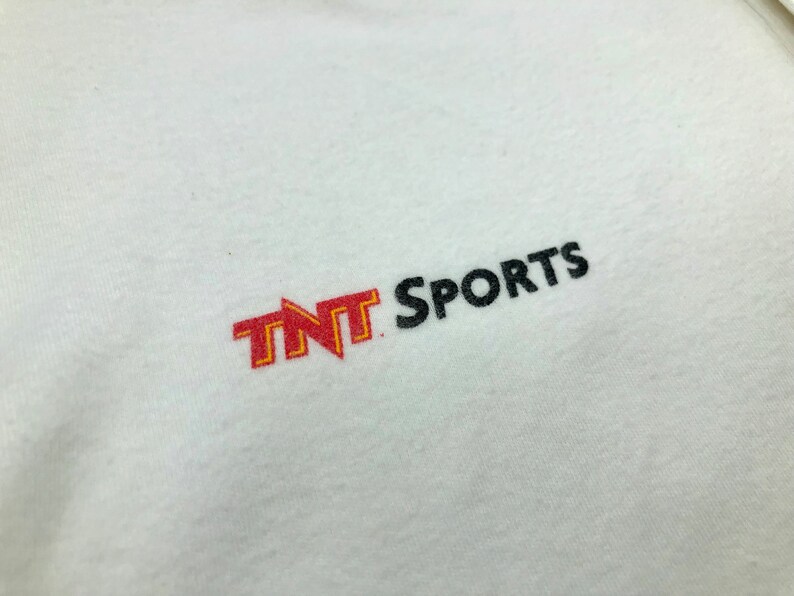 Pu&ograve; includere: Maglietta bianca con un logo "TNT Sports" ricamato in rosso e giallo sul davanti.