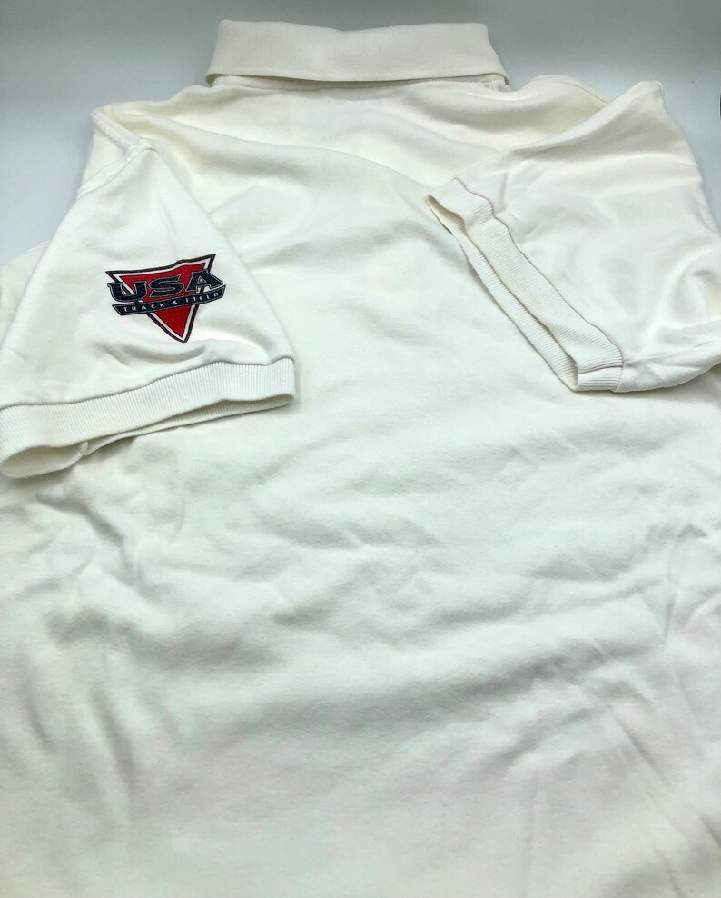 Pu&ograve; includere: Polo bianco con un logo USA Track & Field rosso, bianco e blu sulla manica sinistra.