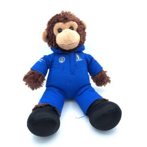 May include: A plush monkey toy wearing a blue astronaut jumpsuit with patches. The monkey has brown fur on its head, arms, and feet, with a tan face and black feet. The toy is sitting against a white background.