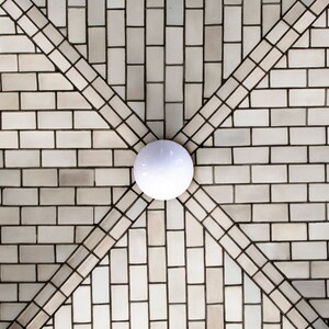 Puede incluir: Una lámpara de techo blanca con una pantalla redonda de cerámica blanca, centrada en un techo de azulejos blancos con un patrón geométrico.