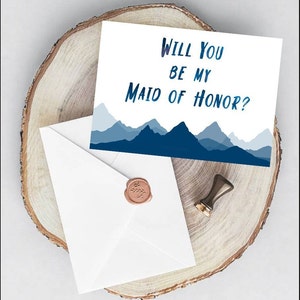 May include: A white card with blue mountains in the background. The text on the card reads "Will you be my Maid of Honor?"