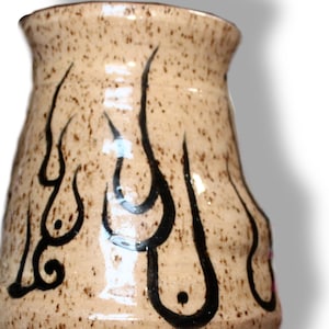 Puede incluir: Un jarrón de cerámica beige moteado con una parte superior acampanada y una sección media bulbosa. El jarrón está adornado con diseños negros en forma de llama. La cerámica hecha a mano tiene un acabado brillante y una estética rústica.