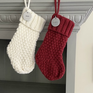 May include: Two knitted Christmas stockings with personalized metal tags. The stocking on the left is white and the stocking on the right is red.