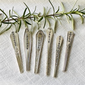 May include: Six silver spoons with black lettering, each labeled with a different type of cheese: Gruyere, Cheddar, Gouda, Brie, Asiago, and Swiss.