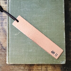 May include: A copper bookmark with a hammered finish and the text "###" engraved on it. The bookmark is attached to a black leather cord.