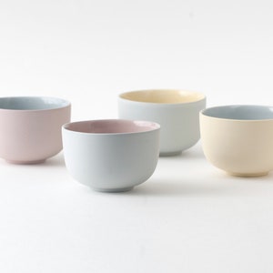 Op de afbeelding: Vier keramische kommen in verschillende pastelkleuren: lichtroze, lichtblauw, lichtgeel en donkerblauw. De kommen zijn allemaal even groot en hebben dezelfde vorm en zijn in een rij op een wit oppervlak gerangschikt.