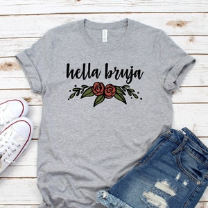 May include: A grey t-shirt with the text "hella bruja" in black lettering and a floral design featuring red roses. The shirt is laid flat on a white wooden surface with a pair of white trainers and denim shorts.