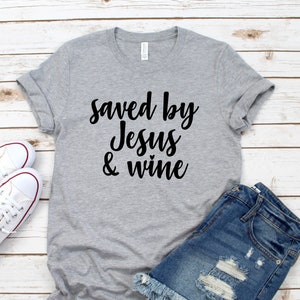 以下が含まれることがあります： 白い木製の表面に置かれたグレーのTシャツ。黒い文字で「Saved by Jesus & wine」と書かれている。白いスニーカーとデニムのショートパンツが一緒に置かれている。