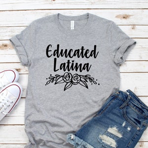 K&ouml;nnte beinhalten: Ein graues T-Shirt mit dem Text "Educated Latina" in schwarzen Buchstaben und einem floralen Design darunter. Das Shirt liegt flach auf einer wei&szlig;en Holzoberfl&auml;che mit einem Paar wei&szlig;en Turnschuhen und einer Jeans-Shorts.