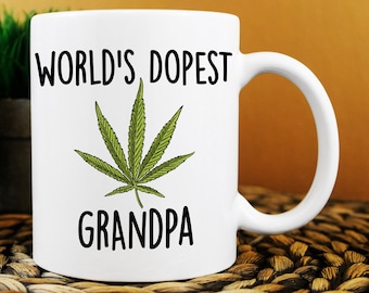 May include: White ceramic mug with the text "WORLD'S DOPEST GRANDPA" in black, featuring a green cannabis leaf illustration. The mug has a curved handle and is set against a neutral background.