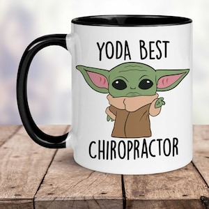 May include: White ceramic mug with a black rim and handle. The mug features a cartoon image of a green alien character with large ears and the text "YODA BEST CHIROPRACTOR".