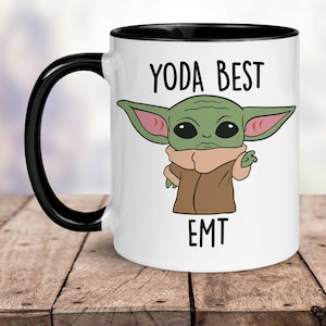 Op de afbeelding: Witte keramische koffiemok met een zwarte rand en handvat. De mok heeft een cartoonafbeelding van een groen buitenaards wezen met grote oren en de tekst "YODA BEST EMT".