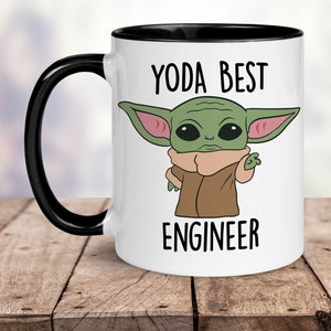 Op de afbeelding: Witte keramische mok met een zwarte rand en handvat. De mok heeft een cartoonafbeelding van een groen buitenaards wezen met grote oren en de tekst "YODA BEST ENGINEER".