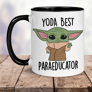 K&ouml;nnte beinhalten: Wei&szlig;e Keramik-Kaffeetasse mit schwarzem Rand und Henkel. Die Tasse zeigt ein Cartoon-Bild eines gr&uuml;nen Au&szlig;erirdischen mit gro&szlig;en Ohren und einem braunen Mantel. Der Text "YODA BEST PARAEDUCATOR" ist auf die Tasse gedruckt.
