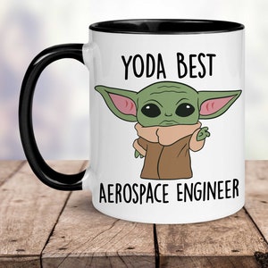 May include: White ceramic mug with a black rim and handle. The mug features a cartoon image of a green alien character with large ears and the text "YODA BEST AEROSPACE ENGINEER".