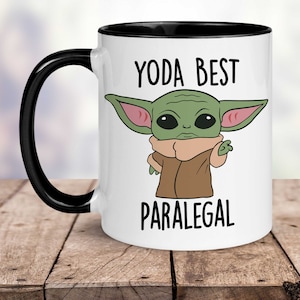 May include: White ceramic coffee mug with a black rim and handle. The mug features a cartoon image of a green alien character with large ears and the text "Yoda Best Paralegal".