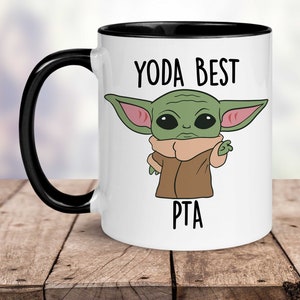 May include: White ceramic mug with a black rim and handle. The mug features a cartoon image of a green baby Yoda character with the text "Yoda Best PTA" printed on the mug.