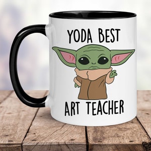 Puede incluir: Taza de caf&eacute; de cer&aacute;mica blanca con borde y asa negros. La taza presenta una imagen de dibujos animados de un personaje alien&iacute;gena verde con orejas grandes y el texto "Yoda Best Art Teacher".