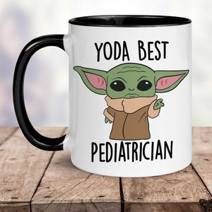 Puede incluir: Taza de cer&aacute;mica blanca con borde y asa negros. La taza presenta una imagen de dibujos animados de un personaje alien&iacute;gena verde con orejas grandes y el texto "Yoda Best Pediatrician".