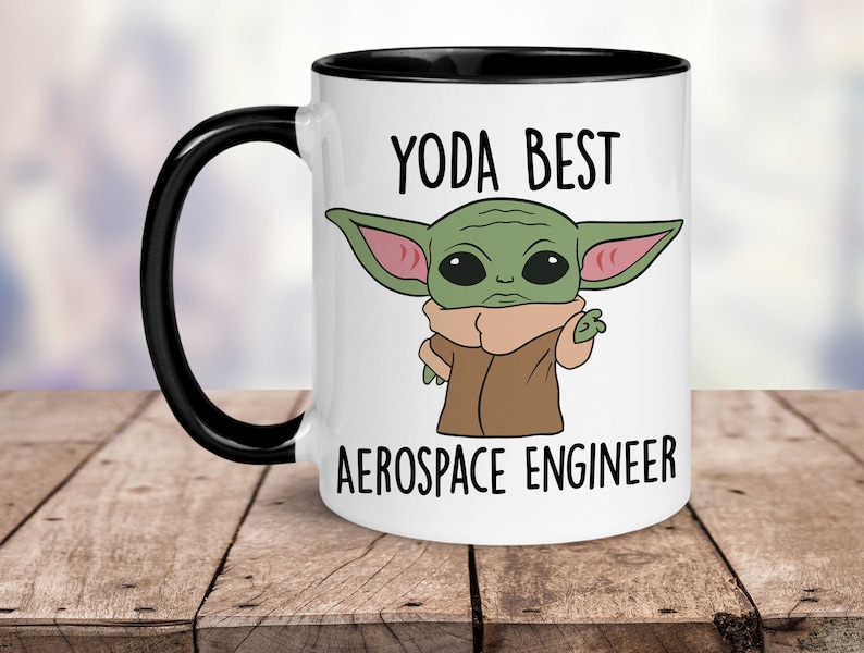 May include: White ceramic mug with a black rim and handle. The mug features a cartoon image of a green alien character with large ears and the text "YODA BEST AEROSPACE ENGINEER".