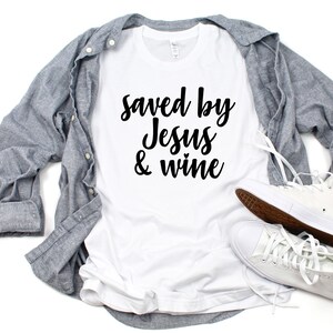 以下が含まれることがあります： 白いTシャツに黒い文字で「Saved by Jesus & wine」と書いてあります。Tシャツは白い表面に置かれ、グレーのボタンダウンシャツと白いハイカットスニーカーが置かれています。