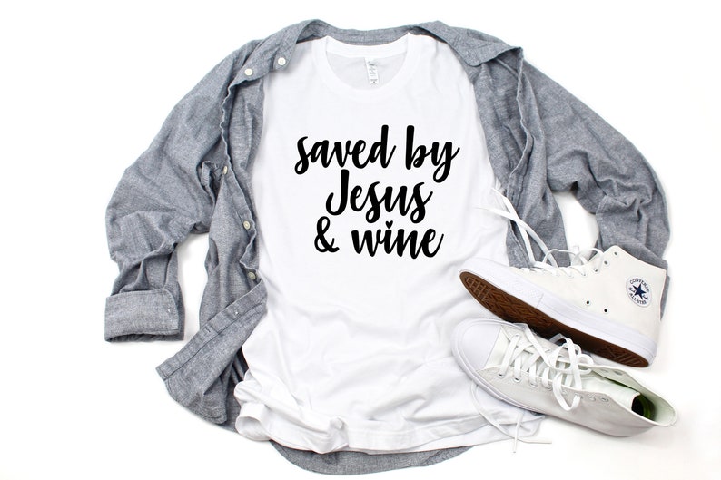 以下が含まれることがあります： 白いTシャツに黒い文字で「Saved by Jesus & wine」と書いてあります。Tシャツは白い表面に置かれ、グレーのボタンダウンシャツと白いハイカットスニーカーが置かれています。