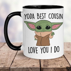 May include: White ceramic mug with a black rim and handle. The mug features a cartoon image of a green alien character with large ears and the text "YODA BEST COUSIN LOVE YOU I DO".