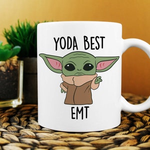 Op de afbeelding: Witte keramische mok met een groen cartoonfiguur en de tekst "YODA BEST EMT".