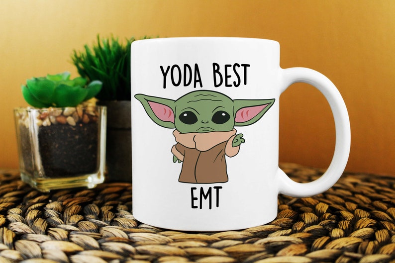 Op de afbeelding: Witte keramische mok met een groen cartoonfiguur en de tekst "YODA BEST EMT".