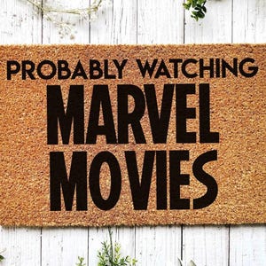 K&ouml;nnte beinhalten: Eine braune Kokosfaser-Fu&szlig;matte mit dem Text "PROBABLY WATCHING MARVEL MOVIES" in schwarzen Buchstaben.