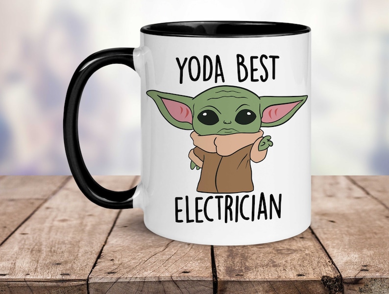 May include: White ceramic mug with a black handle and interior. The mug features a cartoon image of a green alien character with the text "YODA BEST ELECTRICIAN" in black capital letters. The mug is sitting on a wooden surface.
