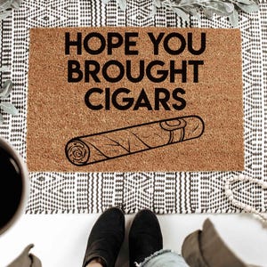 K&ouml;nnte beinhalten: Eine braune Fu&szlig;matte mit dem Text "HOPE YOU BROUGHT CIGARS" in schwarzen Buchstaben. Unter dem Text befindet sich eine Schwarzwei&szlig;illustration einer Zigarre.