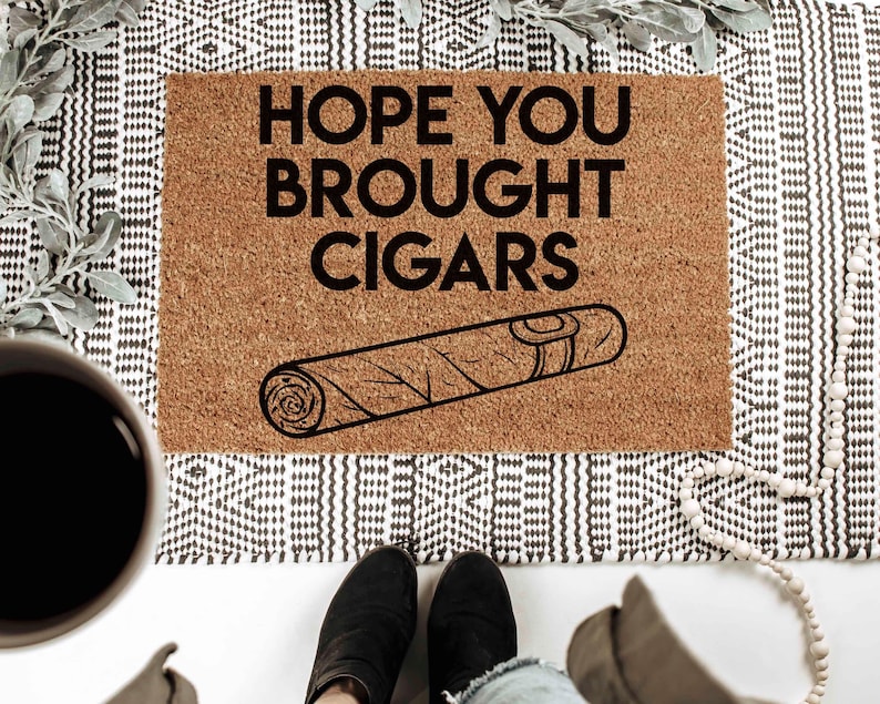 K&ouml;nnte beinhalten: Eine braune Fu&szlig;matte mit dem Text "HOPE YOU BROUGHT CIGARS" in schwarzen Buchstaben. Unter dem Text befindet sich eine Schwarzwei&szlig;illustration einer Zigarre.