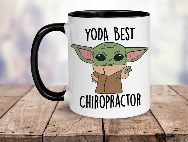 May include: White ceramic mug with a black rim and handle. The mug features a cartoon image of a green alien character with large ears and the text "YODA BEST CHIROPRACTOR".