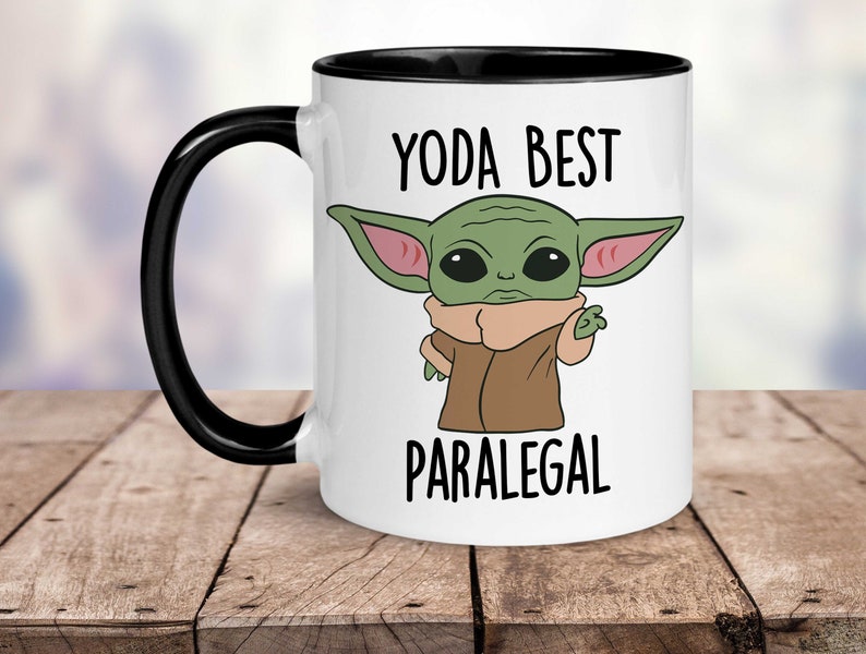 May include: White ceramic coffee mug with a black rim and handle. The mug features a cartoon image of a green alien character with large ears and the text "Yoda Best Paralegal".