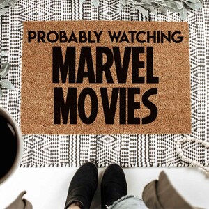 K&ouml;nnte beinhalten: Eine braune Kokosmatte mit dem Text "Probably Watching Marvel Movies" in schwarzen Buchstaben.