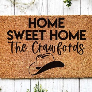 May include: A brown coir doormat with the text "Home Sweet Home The Crawfords" in black lettering. A black cowboy hat is below the text.