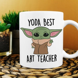 Puede incluir: Taza de cer&aacute;mica blanca con un personaje de dibujos animados verde con orejas grandes y el texto "YODA BEST ART TEACHER" impreso en ella.